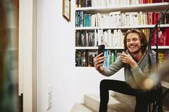 Smiling man sitting on stairs, making a selfie. bookshelf in background