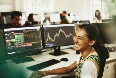 Woman smiling while sitting behind her desk, screens displaying financial information.