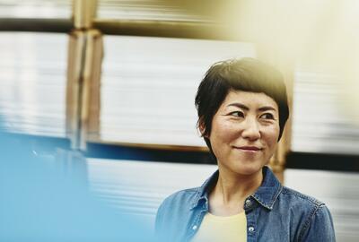 Female Asian worker in logistics environment. Blue-collar. Smiling. Primary color blue. Secondary color white/cream.